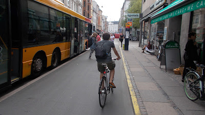 A cyclist on a wide cycle track stops as people get off a yellow bus to his left and cross the cycle track to the footway to the right where there is a bus stop flag.