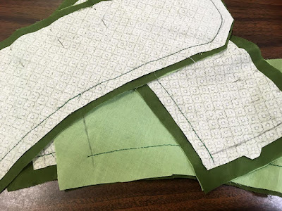 A small stack of narrow curved panels of various green fabrics, pinned along one long edge and stitched in dark green on the other.