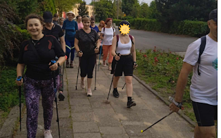 Grupa osób idzie chodnikiem z kijami, uprawiają nordic walking.