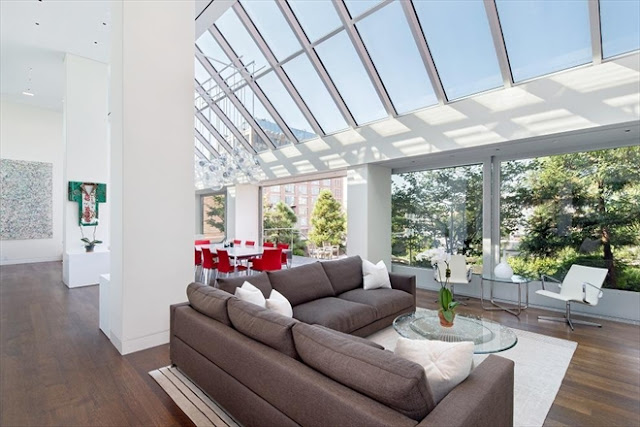 Tribeca penthouse with wood floors, a brown sofa with white accent pillows facing a large windows with a view of green plants under a glass ceiling 