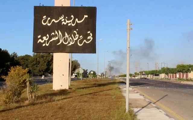   Image Attribute: A sign which reads in Arabic, "The city of Sirte, under the shadow of Sharia" is seen as smoke rises in the background while forces aligned with Libya's new unity government advance on the eastern and southern outskirts of the Islamic State stronghold of Sirte, in this still image taken from video on June 9, 2016. Reuters TV/File Photo