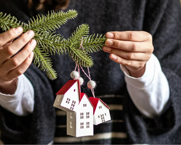 hands holding evergreen branch on which three white paper houses with red roofs are hanging