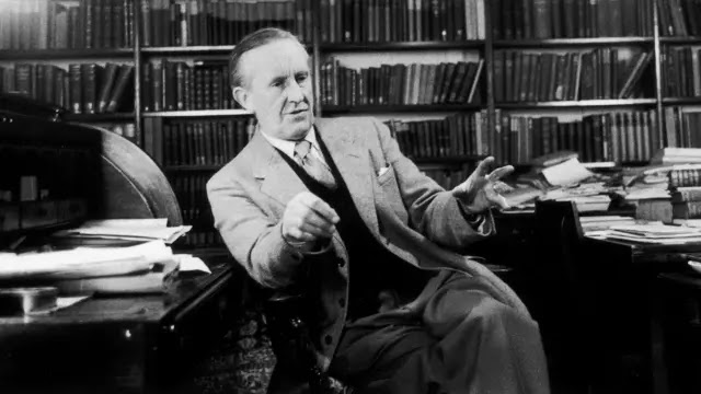 Foto clássica de Tolkien onde vemos o escritor fumando seu cachimbo enquanto parece falar alguma coisa. Ele é um homem branco, idoso, está de terno e ao fundo temos prateleiras cheias de livros.