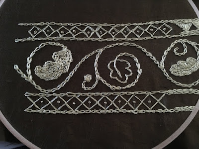 An embroidery sampler in gold thread on olive-green silk stretched in a wooden embroidery hoop, showing some narrow borders with diamond fill, with a large scrolling vine down the center panel. The vine carries three oak leaves, one outlined, one filled with chain stitch, and one filled with satin stitch, and a single acorn..