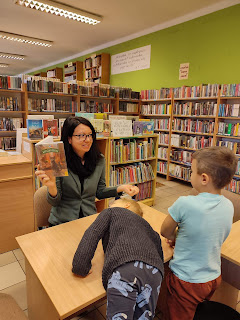 Pani bibliotekarka opowiada dzieciom o przeczytanej książce. W prawej ręce unosi książkę do góry. tło: Sala biblioteczna.