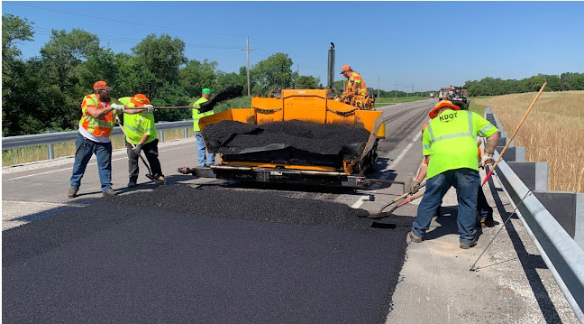 Four KDOT crew members spread black asphalt on a road with equipment