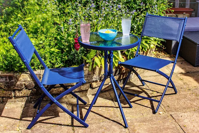 2 blue fold up chairs and a small table in a garden in front of a flower bed