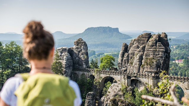 Das Foto zeigt die Basteibrücke in der Sächsischen Schweiz. Man hat einen wahnsinnig schönen Blick auf die Berge und Wälder. Verschwommen im Vordergrund erkennt man die Rückenansicht einer jungen Frau mit einem weißen Shirt und einem gelben Rucksack. Sie ist auf Wanderschaft. bei leichtbewölktem Wetter.