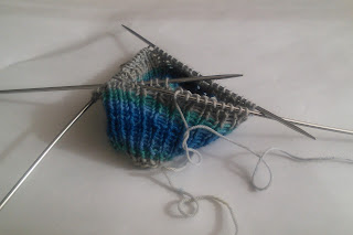 A ribbed sock cuff on double-pointed needles.  The yarn stripes from cream to blue, grey, teal, blue, and cream. 