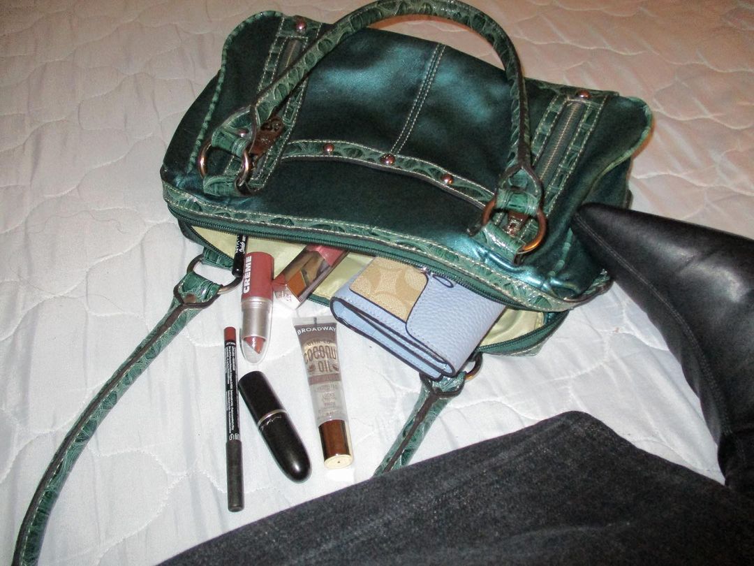 Green large hobo bag with long straps, blue Coach wallet, clear lipgloss, MAC lipstick on white bedspread with women's leg wearing blue flared jeans and black pointy toe spike heel boots