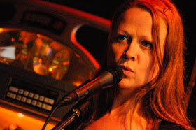 Photo of poet Jeanann Verlee speaking into a microphone. She has long, reddish hair. There is a jukebox in the background.