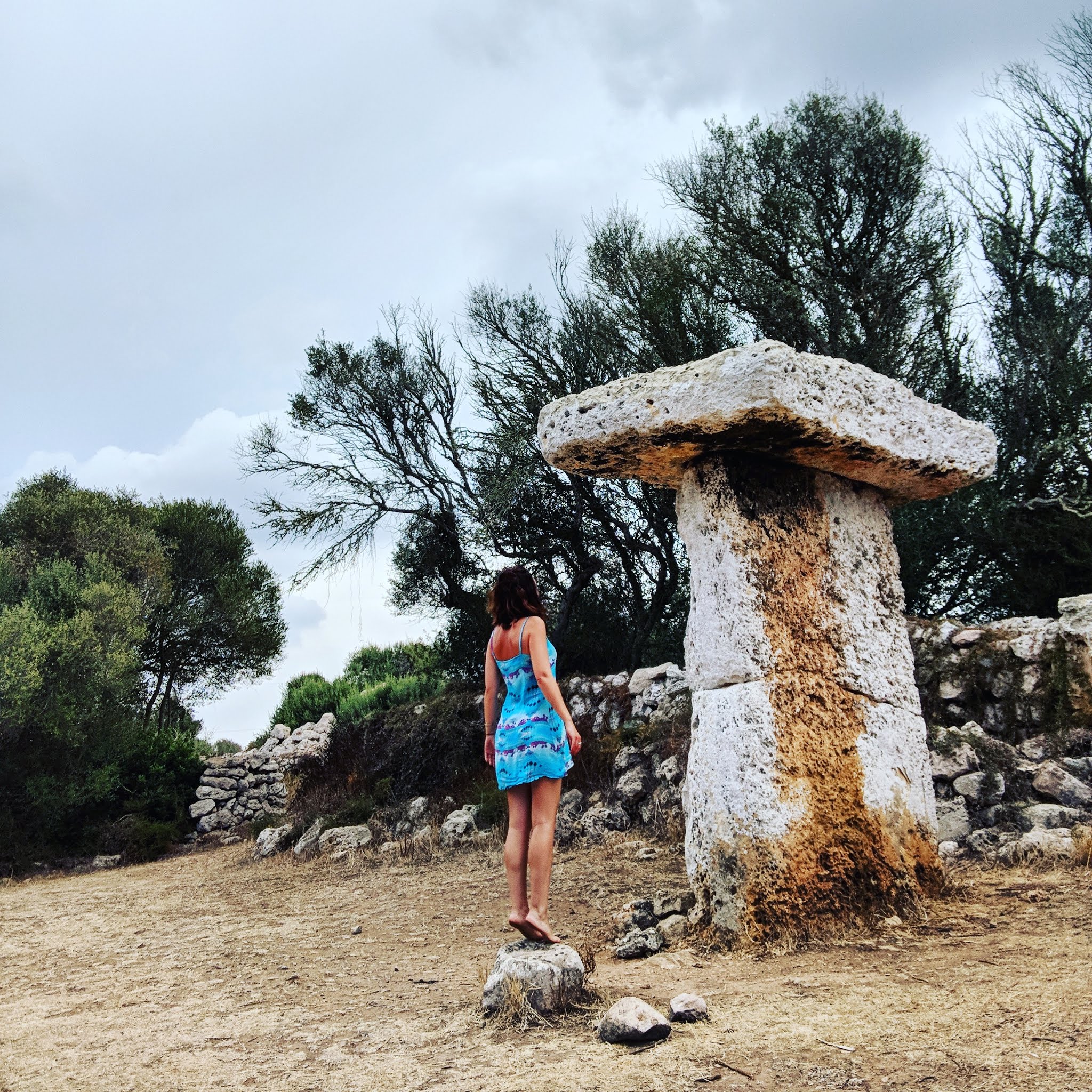 Lydia swinscoe wearing a short blue summer dress standing on tiptoes admiring the ancient stone structure known as a taula on the island of menorca