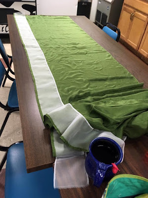 A long panel of green silk laid out on a brown plasticky break room table, with the excess piled up at the near end, and a narrow strip of hemmed white organza laid just along the left edge.
