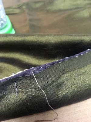 A close-up of the upturned edges of two pieces of fabric, showing small tan running stitches wandering off the edge of he lower, green-gold fabric and only following the turned-down edge of the brown fabric.