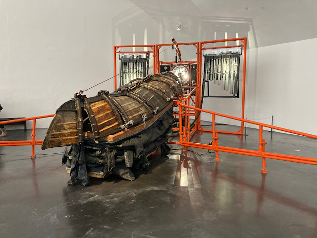 A large sculpture with an orange metal frame and an upside down wooden boat.