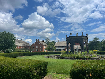 The Rapp House garden at Old Economy Village with a brilliant blue sky and puffy white clouds. Green hedges and grass in the foreground and the stone six-sided pavilion towards the right. The pavilion has a white statue in the center and is surrounded by a circle of foot-high flowering plants and then by water. The George and Frederick Rapp Houses as well as the Feast Hall are visible beyond the grass and pavilion.