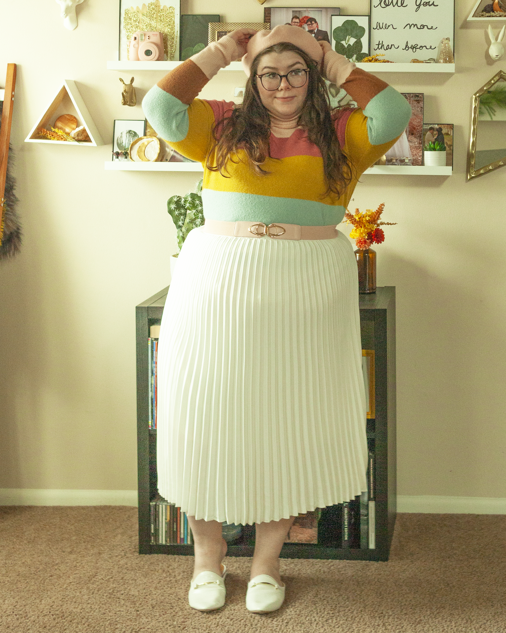 An outfit consisting of a pastel pink beret, a light brown, pastel pink, pastel blue and marigold yellow turtleneck sweater tucked into a white pleated midi skirt, belted with a pastel pink belt and white pointed toe loafer inspired mules.