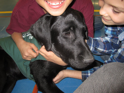 A blissful black lab puppy Romero is being hugged by two young boys in a school gymnasium. Romero is wearing his green future dog guide jacket. The boy behind Romero is wearing a maroon t-shirt and has his arms wrapped around Romero's neck. His face is cropped out of the picture but you can see his huge smile, completely with newly grown-in front teeth! The other boy is on the right side of the picture at Romero's head. He is wearing a blue plaid shirt and scratching under Romero's chin and chest. He is gazing down lovingly at Romero's face.