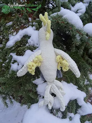 Na drzewie iglastym posypanym śniegiem siedzi odwrócona tyłem wykonana na szydełku z pluszowej włóczki kakadu żółtoczuba