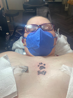 A selfie taken by a brunette, bespectacled, middle aged white woman wearing a blue FFP2 mask, laying on her back on a tattooist's bed. She's topless apart from nipple covers made out of kitchen towel that are taped in place. She has a tattoo over her heart of a cat's paw print, and the name 'Betty' below the paw print. The tattoo is black, and the skin around the tattoo is red where it's literally just been tattooed.