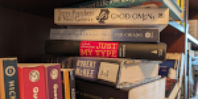 A pixelized view of a bookshelf which is full, poorly organized, and about the only easily made out thing is Good Omens by Neil Gaiman.