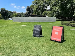 Green grass lawn leading up to a grey concrete wall. Two sandwich board signs are shown one in black and the other in dark orange.