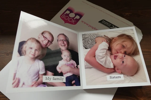 The first 2 pages of the photo book showing a family portrait called "my family" and 2 girls hugging called "sisters" book rests on top of envelope with the POP BOOK image showing