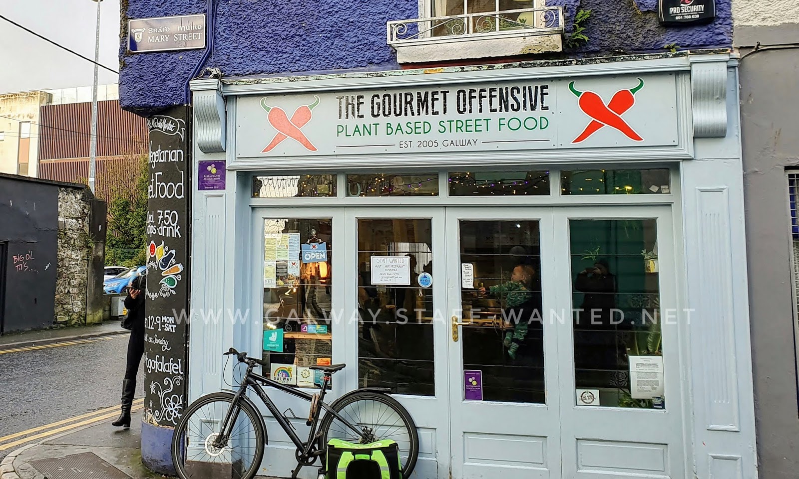 Restaurant shopfront and door, with job advertisement sign on the window and a Deliveroo rider's bicycle dumped immediately outside the door while the rider collects an order.