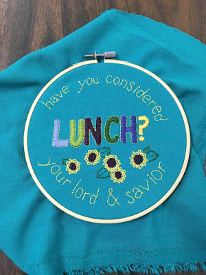 A plain wooden embroidery hoop with a dull silver screw, holding a orugh square of turquoise fabric embroidered with "Have you considered your lord and savior" in small yellow backstitch around the edges of the circle, and "lunch" in large satin-stitch capital letters of lavender, two shades of green, garnet, and navy. "Lunch" ends in a bright-yellow question mark, and five scattered sunflowers with outlined leaves fill the space below.