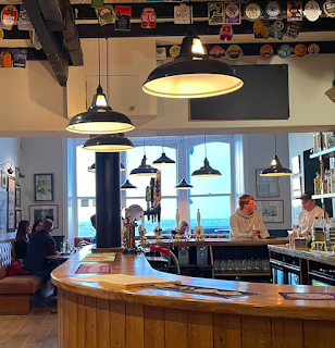 A photo of a pub, with a bar, and a large window in the back ground. You can see the sea and the sky through it, and they're both a clear, bright blue.