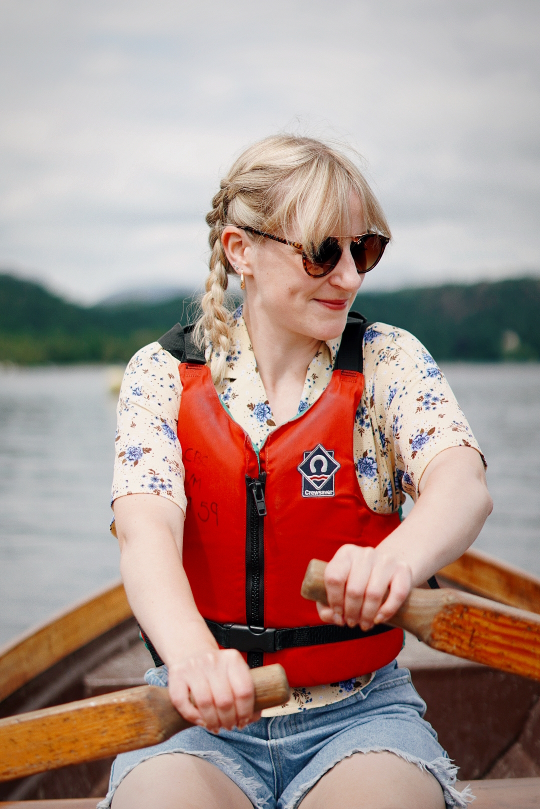 Amy is sat in a rowing boat, oars in hand. Behind is a big lake. She is wearing a bright red life jacket.