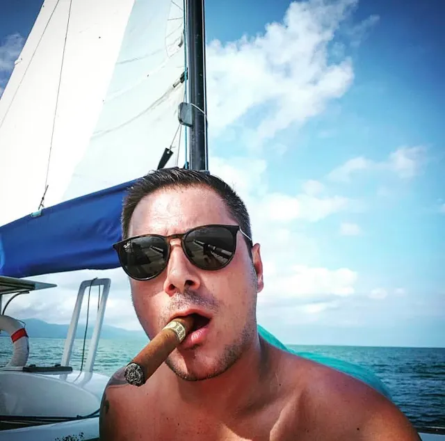 From shoulders up dark-haired man wearing sunglasses shirtless with a cigar in his mouth taking a selfie