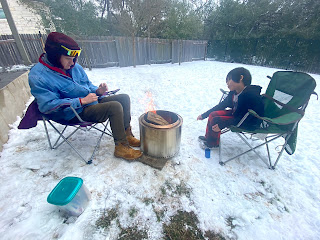 TJ, wearing Timbs, several jacket layers, a winter hat with earflaps, and reflective sunglasses, sits in a camp chair across from Tian, wearing plaid pajamas and several jackets. They are outside with snow covering the ground. Between them is a silver round camp stove with a blazing fire.