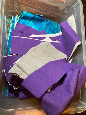 A pile of carefully cut fabric in a clear container. Some of it is gray, other pieces are purple, and towards the bottom are pieces of a swirling blue and green fabric.