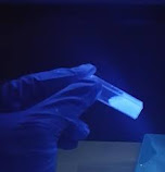 The image shows a scientist holding a test tube with a glowing liquid. The liquid contains per- and polyfluoroalkyl substances (PFAS), also known as "permanent chemicals". These chemicals are used in many products, including non-stick pans, waterproof clothing and fire extinguishers.