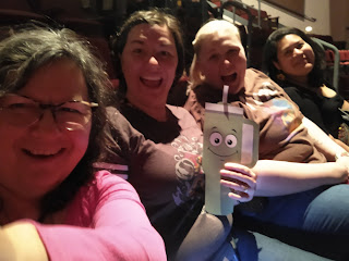 A slightly blurry selfie of four women and a flat cartoon drink cup named Flat Stanley. They are laughing and smiling and sitting in theater seats next to each other.