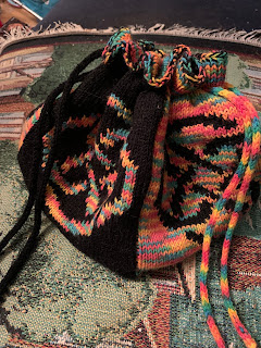 A handknit drawstring bag. The sides are colorblocked in alternating colors: a black background with a rainbow knotwork motif, next to a rainbow background with a black knotwork motif. The drawstrings are i-cords, one black and one rainbow.