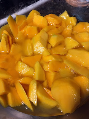 A close-up of large chunks of fresh mango partially submerged in clear liquid, in a clear glass bowl.