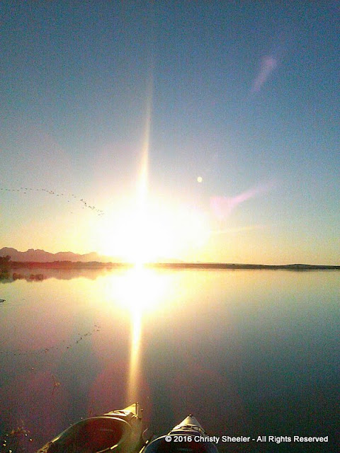 As sun sets low in the sky, its bright light reflects of the water.  Two kayaks in the foreground.