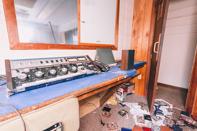 The corner of a defunct radio station studio, featuring an aged mixing console strewn with dust and a few scattered items, including a telephone and speaker. The floor is cluttered with discarded cassette tapes, hinting at the hasty departure from a once active broadcasting hub.