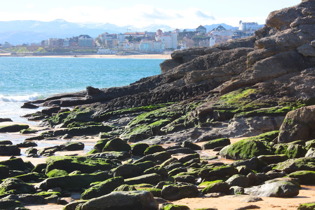 Widok na dzielnicę Sardinero w Santander z plaży Los Molinucos. Za niewielką plażą, zarzuconą kamieniami farbowanymi algami na zielono falują spokojnie wody zatoki. Na przeciwległym brzegu widoczne jest pasmo piaszczystej plaży i wznoszące się nad nią na wzgórzu budynki dzielnicy Sardinero. W tle widać ośnieżone górskie szczyty, nad którymi zbierają się deszczowe chmury.