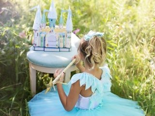 A girl in a blue princess dress holds a star wand, with a castle toy in front of her, engaged in imaginative play.