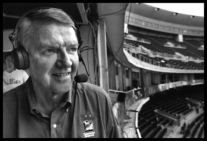 A close-up of Harry Kalas wearing headphones in the broadcast booth of a stadium, looking out at the field