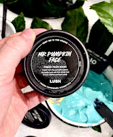 A photo of a white hand holding a circular black tub filled with scrubby orange face mask with a circular black label that says mr. Pumpkin face fresh face mask in white font on a bright background