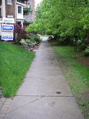Sidewalk with house porchsteps on the left and grassy lawn on the right, with trees providing canopy cover (west side of Queen Elizabeth Drive near McLeod Street)