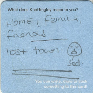 Pale blue beermat with handwritten text that reads "Home, family, friends. Lost town." There is a small drawing of a sad face in the bottom right corner, which has been labelled with the word '"sad".