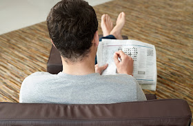 Young man sitting on sofa doing crossword puzzle in newspaper back view