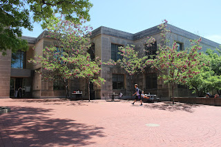 The entrance to the library, taken on a bright sunny day. The building has an almost jagged skyline as it is built in 4 slightly offset blocks. A student is walking across the center of the photo. The image is called the Speakers Circle entrance because the pavement leading to the entry is made of brick in a circular pattern.