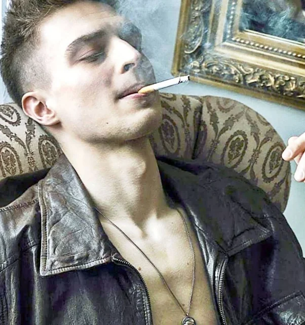 Man wearing a leather jacket with no shirt on underneath smoking a cigarette from the chest up