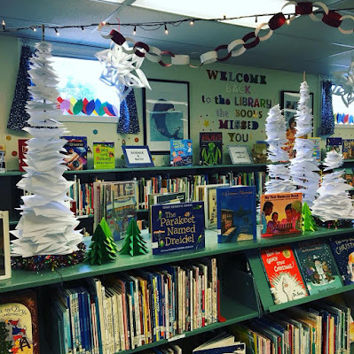 View of bookshelves and counters in library. White holiday lights are strung across ceiling, and pieces of paper are arranged on tall spindles, graded from larger sheets at base, to smaller at-top to suggest snow-covered trees. Winter holiday themed books are displayed on counter-top.